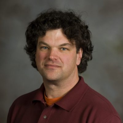 dr. jay wilkins looks into the camera in this formal portrait. he wears a maroon one-quarter buttoned pullover with an orange shirt underneath