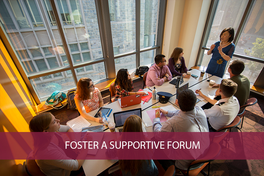 faculty member standing at end of a conference table sharing with a group of students - (text) Foster a Supportive Forum