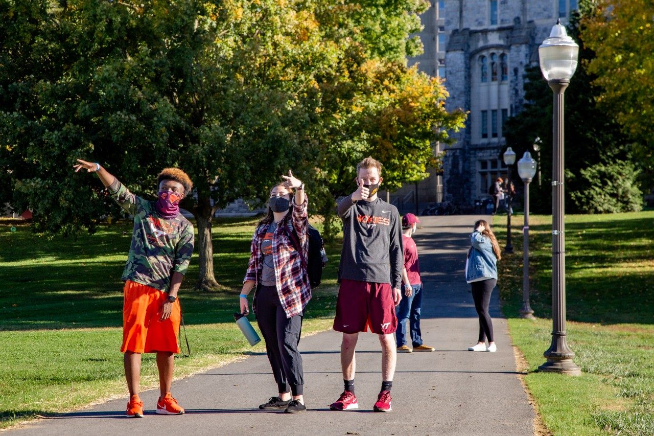 three young adult students happily gesture to the photographer as they walk across campus. They are a back man, a white woman and a white man enjoying themselves