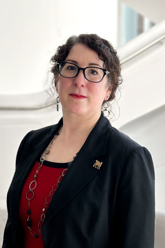 A woman wearing black-rimmed glasses, black jacket, and red shirt.