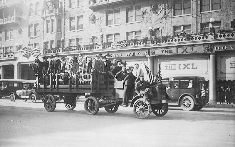 A black and white photo showing citizens and soldiers on a cart rolling down a street in Stockton, California to celebrate the end of the first World War. The cart has several people standing in it, and most are wearing masks to protect against the flu.