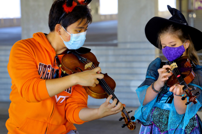 James Kim, a first-year music education student, holds a violin and helps a student play a violin. The young student wore a witch hat and a face covering. Kim wore a Virginia Tech sweatshirt and a face mask. The two both donned Halloween costumes for the String Project gathering in the Perry Street Parking Garage in Blacksburg. The Virginia Tech String Project is a community-outreach program, offering small group and individual instruction on a variety of string instruments. Instruction is provided by graduate and undergraduate teachers, under the supervision of Music faculty