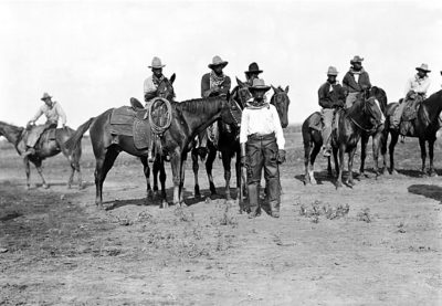 A black and white photograph showing one cowboy standing amongst several on horseback.