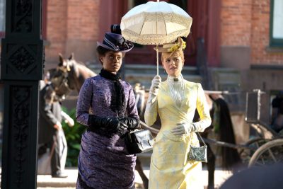 A Black woman and a white woman dressed for life in the 1880s with hats and a parasol.