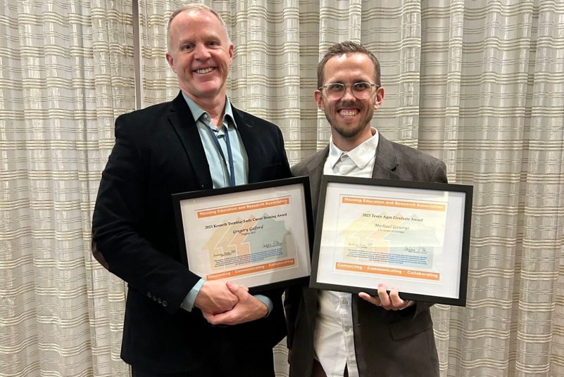 Two men are smiling and standing side by side. They are each holding framed awards.