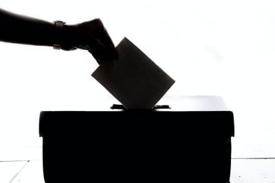 Someone's hand is shown placing a ballot into a voting box 