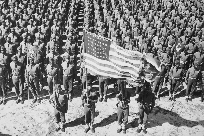 Hundreds of soldiers at Ft. Bragg on parade with one soldier holding a prominent American flag in the foreground.