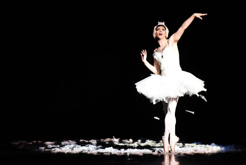 A male ballerina dressed in a white leotard and a feathered tutu poses en pointe on a black stage with a black background. Feathers are falling from the tutu and a pile of feathers surround the dancer on the floor.