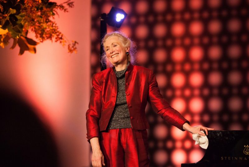 Pianist Barbara Nissman, an older white woman with blonde hair pulled into a bun, wears a red silk suit and grey sweater and stands with one hand on a Steinway piano.