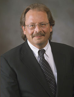 Anthony Colaianne, dressed in a suit and glasses, smiles for a headshot in front of a grey background. 