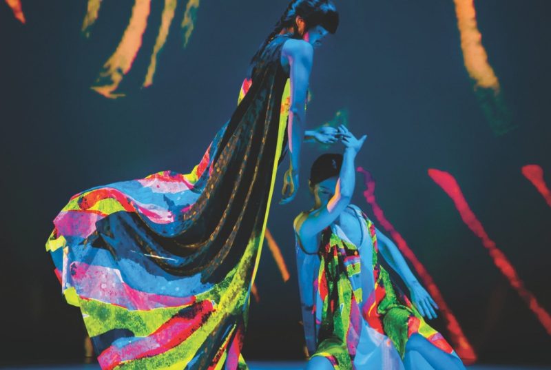 Two dancers with Cloud Gate Dance Theatre of Taiwan perform on stage wearing neon costumes that glow under a black light. One stands over the other, who crouches with one arm in the air. Photo by LEE Chia-yeh