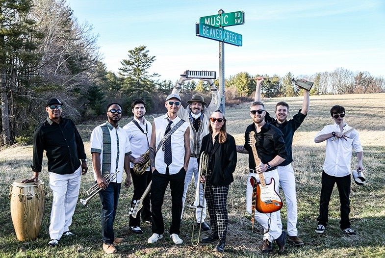 Nine members of Music Road Co band, dressed in black and white, stand outside under street sign, holding various musical instruments