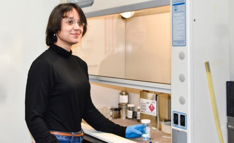 A young women holds a beaker of clear liquid while standing in a lab.