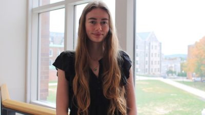 A smiling girl leans against a railing, a big window behind her. 