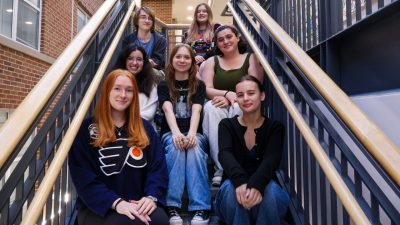 Seven Virginia Tech students, members of the English Club, sit together on an indoor staircase. They are smiling and casually dressed, surrounded by brick walls and railings.
