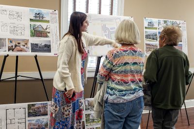 A young woman in a long dress and cardigan points at poster boards on easels as she talks to two women.