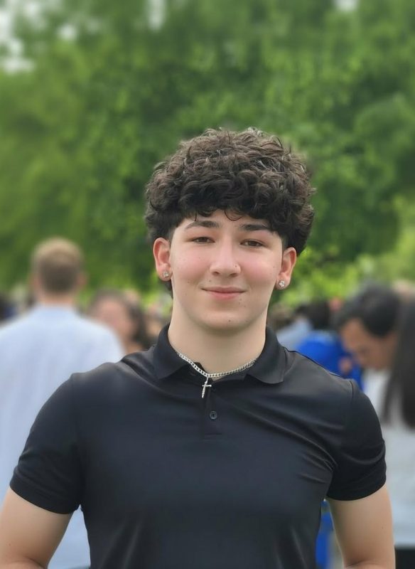 Male with curly hair standing outside with crowd in background.