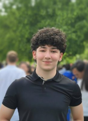 Male with curly hair standing outside with crowd in background.