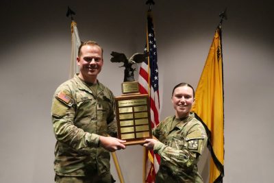 A man and a woman, dressed in Army uniforms, hold a trophy, smiling.