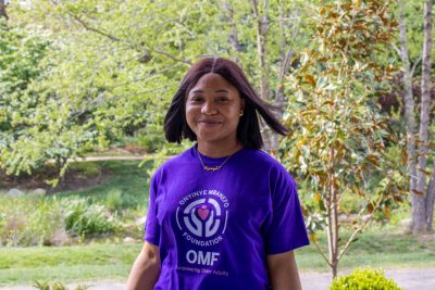A youn woman wearing a bright purple shirt stands in front of several trees, smiling.