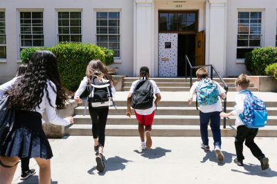 A group of middle-school aged children wearing backpacks run toward their school.