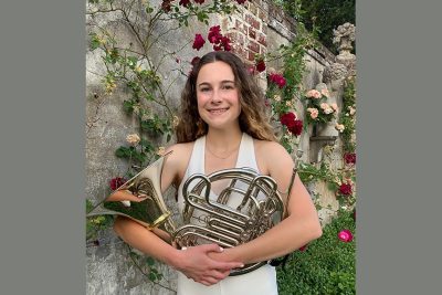Autumn Edson wearing a white halter dress, holding a French horn, standing in front of a wall with roses growing on it.