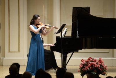A woman in a sleeveless long blue dress stands on stage playing violin with a grand piano and accompanist behind her.