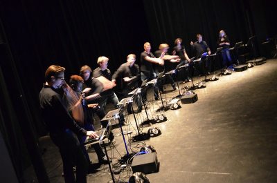 Students in the L2Ork laptop orchestra dressed in black, stand in front of a music stand holding a laptop computer.