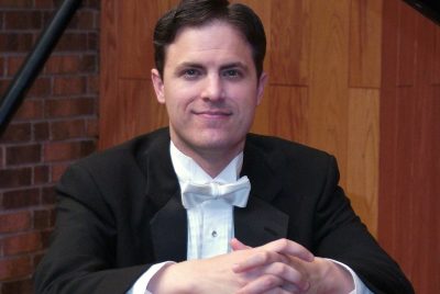 A man dressed in a tuxedo sits at a piano with his hands folded.