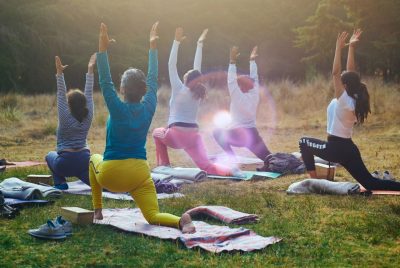 Five people doing yoga outdoors, kneel in a pose on mats with their arms outstretched to the sky.