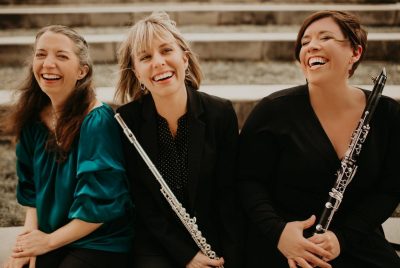 Alma Ensemble, three women sitting outdoors on the steps, laughing, one holding a flute and another holding a clarinet