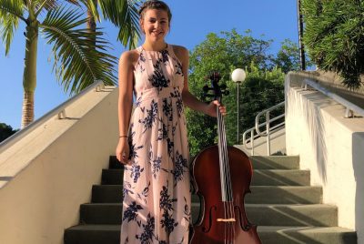 Josie Chase stands outdoors in a sleeveless, long pink dress holding a cello