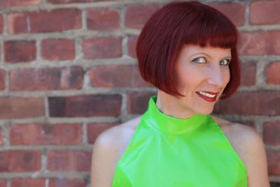 A woman with short red hair, wearing a bright green halter top, in front of a brick wall.
