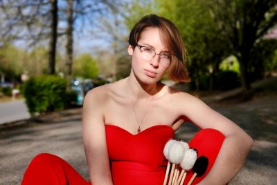 A young woman dressed in a red strapless top and pants, sits on the ground holding percussion mallets.