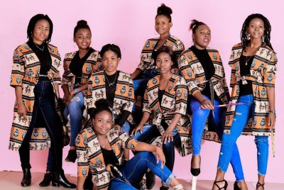 Eight women of the Daughters Band pose against a pink background, wearing blue pants and an orange and black jacket.