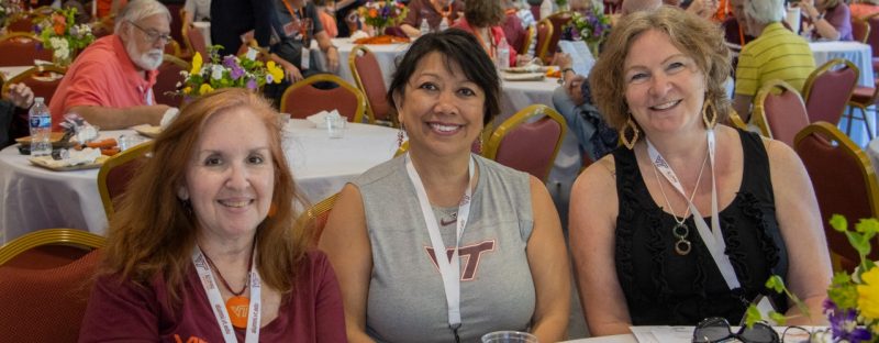 Three women sitting at a table, smiling at a social gathering with other people in the background.