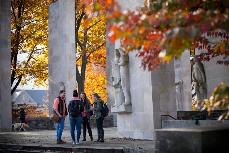 four students stand chatting near the pylons on the blacksburg campus. the leaves are colored in yellows, reds, and oranges. The students wear clothing indicating fall weather.