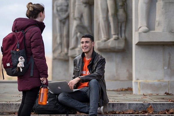 student smiling while seated with laptop at the war memorial talking to another student