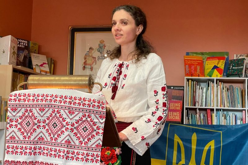 Kira Gulko Mores stands at a podium. She is dressed in a white shirt with embroidered red flowers, traditional Ukrainian clothing.