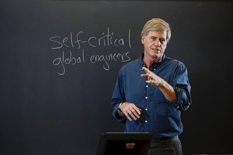 Faculty Lecturing with self-critical global engines written on the a blackboard behind them