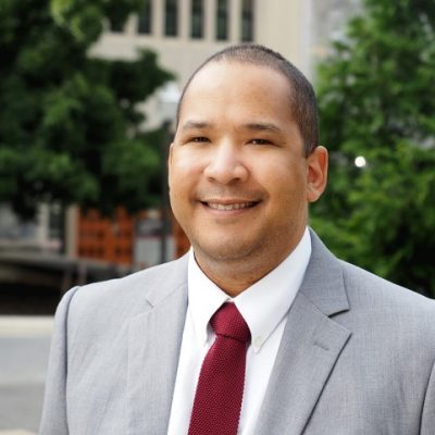 marcus johnson is an african american man - he wears a light grey suit with a starched white button down shirt and a maroon tie. He smiles confidently at the camera.