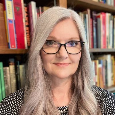 dr. christine christianson smiles calmly at the camera, her face framed by black-rimmed glasses. Her long hair is curled at the ends, and she wears a polka dot dress as she stands in front of bookcases