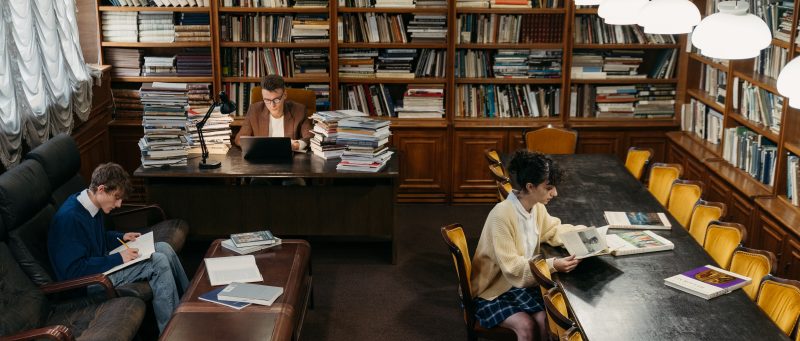 two young white adult men and a young latina adult woman study in a library surrounded by books