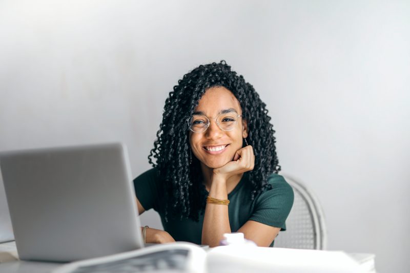 an young african american adult smiles confidently at the camera. she is seated at a table in front of a laptop and has a text book open in front of her. Her hair is curly and shoulder length. She wears a green sweater