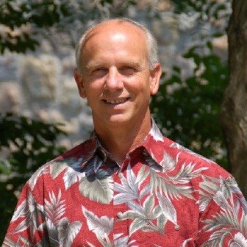 John Wells smiles cheerfully at the camera. He has short cropped grey hair with a bald pate, and is wearing a red hawaiian floral shirt