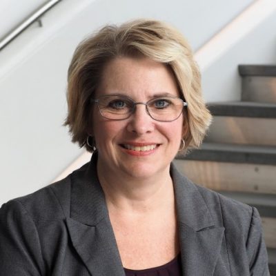 Nancy Bradley, professor in the School of Education, smiles confidently at the camera, wearing a grey suit and a black shirt. Her blonde hair is short and she wears glasses