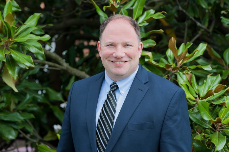 Dr. Gerard Lawson smiles for the camera. he stands in front of greenery, wearing a dark blue suit with a light blue button down shirt and a blue tie with red stripes