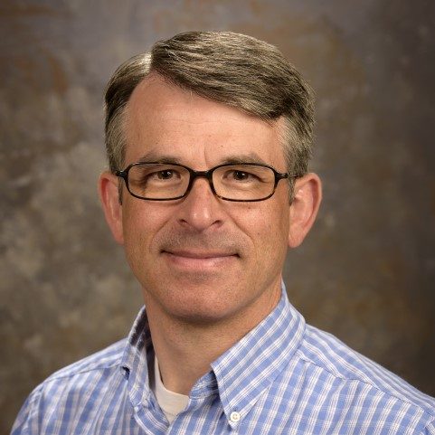 Dr. Dana Robertson, a middle aged white man, wears a blue and white plaid button down shirt and glasses. He smiles for the camera.