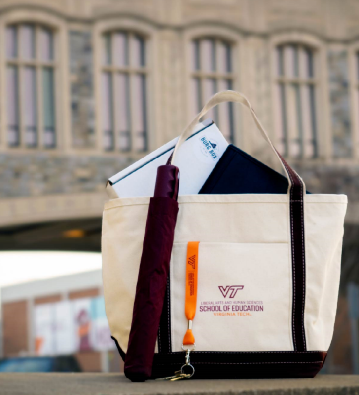 a canvas bag with the school of education sits on wall of hokie stone; we can see a branded maroon umbrella, an orange lanyard keychain, a black leather portfolio, and a box peeking out of the top of the bag