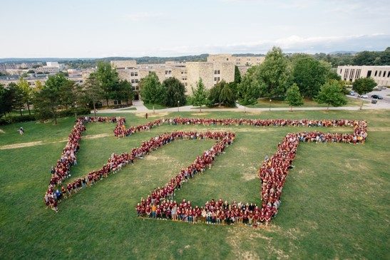 a large group of students, most wearing VT maroon shirts, form a huge VT symbol on the drill field - the image is from above the large group, looking down